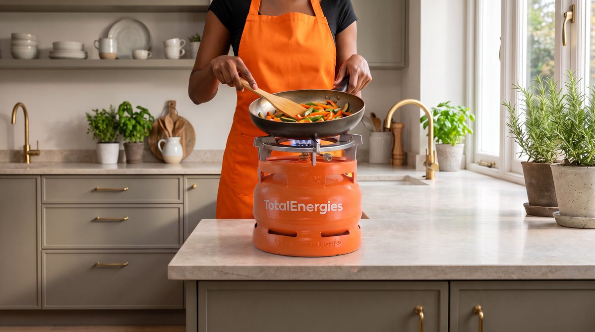 Generated image: a cook stirring vegetables in a pan on a burner mounted on an orange TotalEnergies 6 kg LPG cylinder, on the worktop of a modern kitchen.